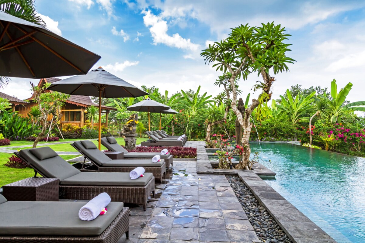 A serene pool area features loungers and umbrellas arranged alongside the tranquil water. Tropical plants and vibrant flowers frame the scene, creating a lush green backdrop. The pool's reflective surface mirrors the surrounding foliage and sky, inviting relaxation.