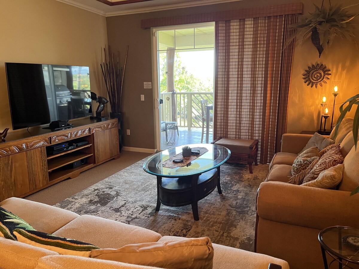 A spacious living area is shown with a large flat-screen TV mounted on a wooden console. Comfortable seating options include a beige sofa and two decorative chairs arranged around a circular glass coffee table. Natural light fills the room through sliding glass doors, leading to a balcony.