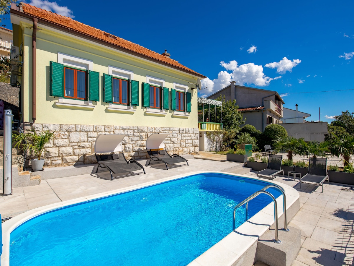 The outdoor pool is surrounded by a tiled terrace featuring two deck chairs. A well-maintained house with bright green shutters is visible in the background, complemented by clear blue skies and scattered clouds.