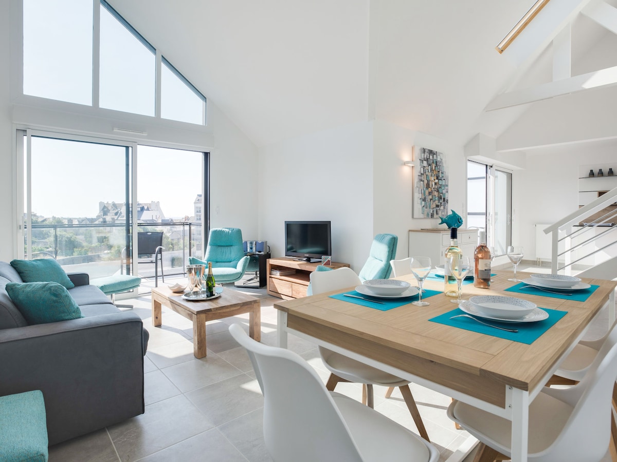The open-plan living area features large windows providing ample natural light. A modern television is positioned beside a cozy seating arrangement. A dining table is set with tableware, and a light-colored couch sits adjacent, enhancing the spacious atmosphere.