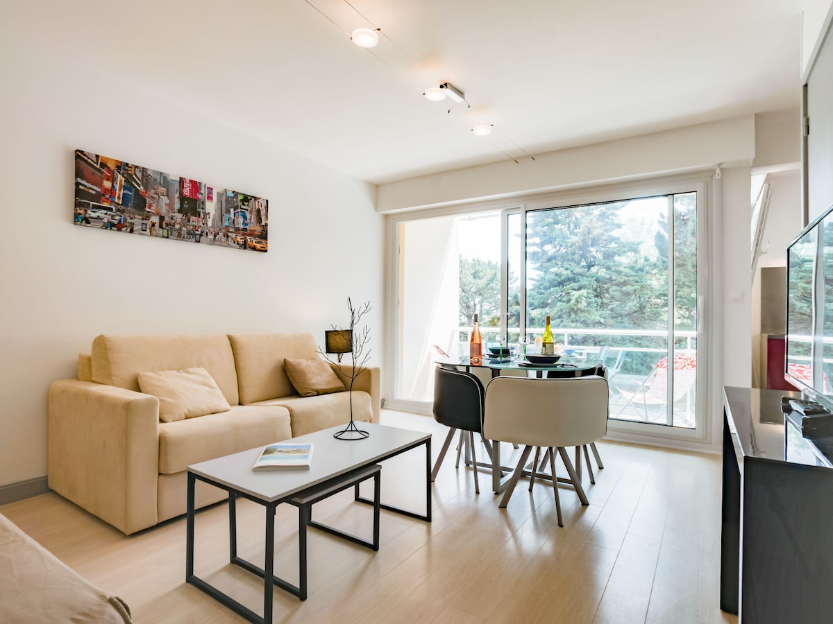 The living area features a comfortable sofa facing a flat-screen TV. A dining table with four chairs is positioned beside a large glass door that opens to a balcony, allowing natural light to fill the space. Decorative artwork adorns the wall.