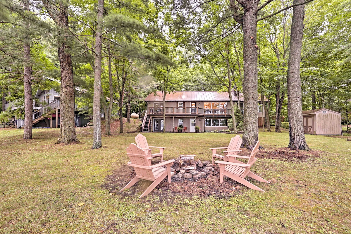 A peaceful outdoor space features four light-colored adirondack chairs arranged around a circular fire pit, surrounded by grass and trees. In the background, a two-story cabin is visible, nestled among the greenery, providing a serene setting for relaxation.