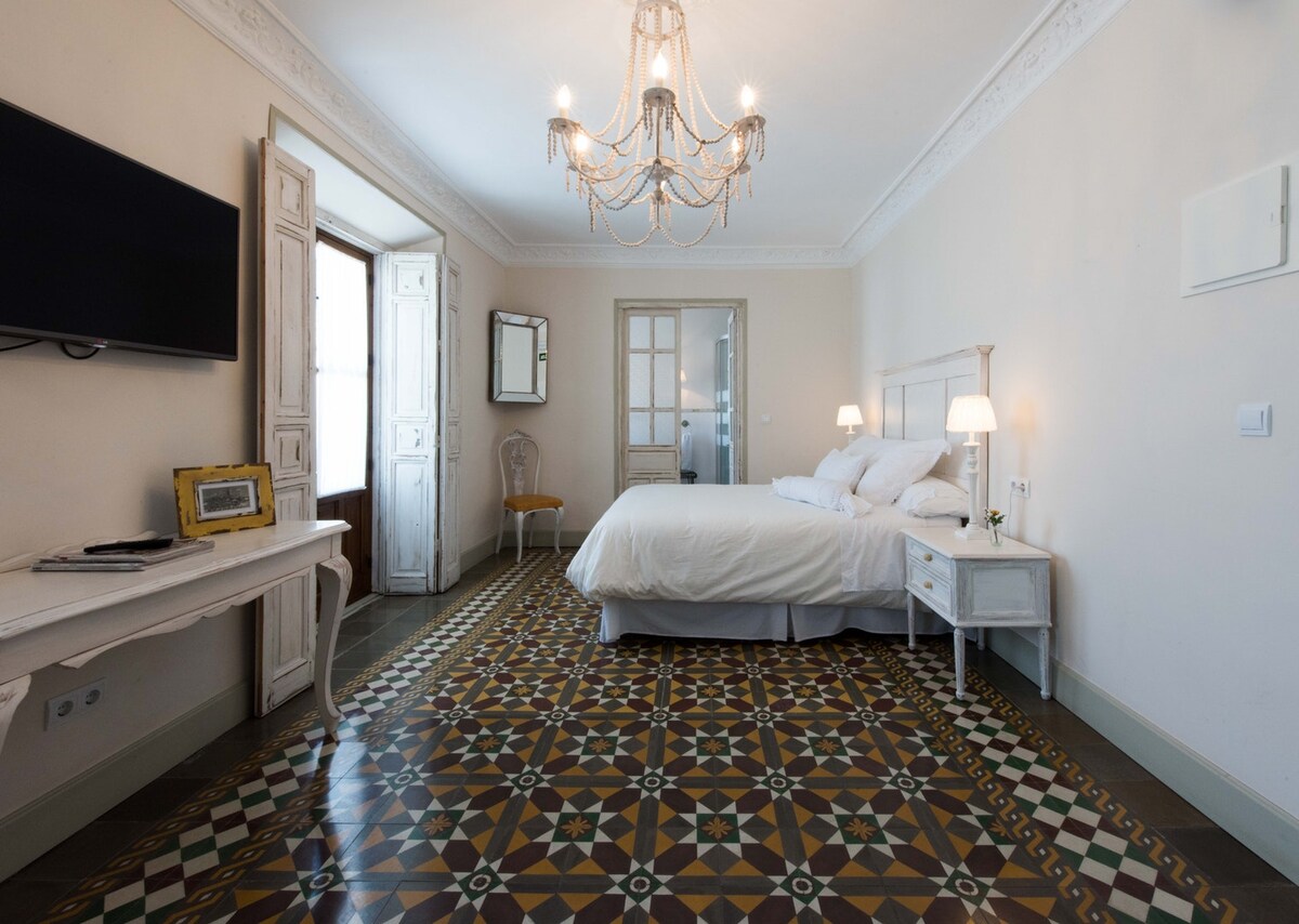 A spacious bedroom features a large, comfortable bed flanked by bedside lamps. A decorative chandelier hangs over the room, while the patterned tile floor adds elegance. Large windows allow natural light to fill the space, complementing the neutral wall tones.