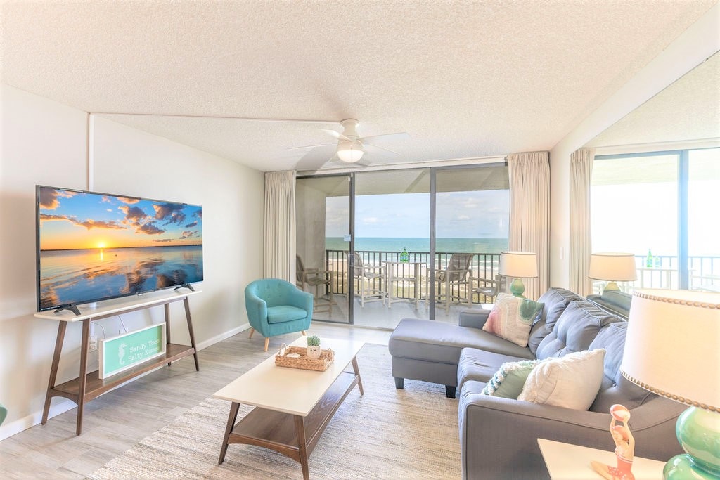 A comfortable living area is presented with a large sectional sofa and a stylish coffee table. A flat-screen TV is positioned on a stand, with wide glass doors providing views of the ocean and access to the private balcony.