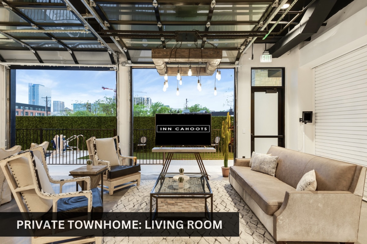 A spacious living room is illuminated by natural light streaming through large glass garage doors that open to a view of the garden. Comfortable seating, including a sleek sofa and several armchairs, is arranged around a central table, offering an inviting space for gatherings.