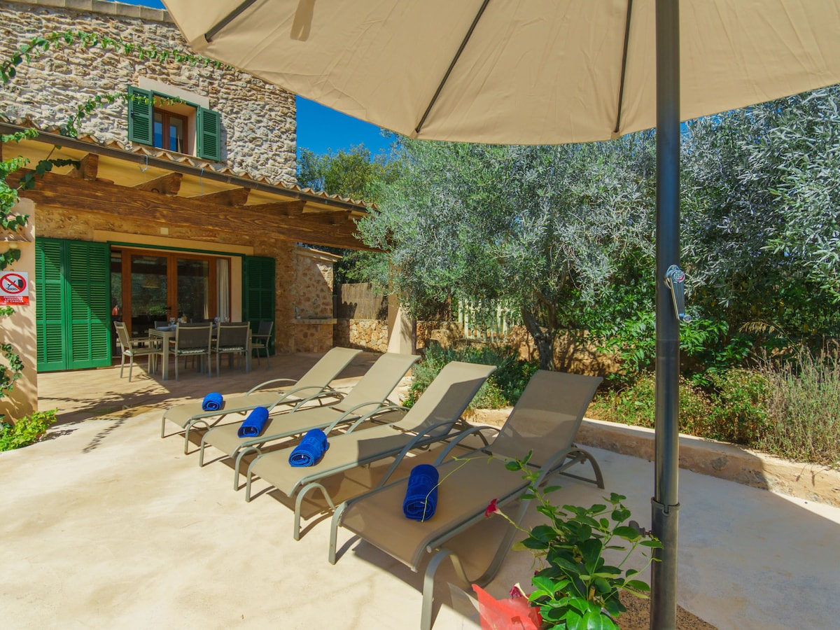 A shaded outdoor area features several sun loungers arranged neatly under an umbrella. The backdrop includes a stone exterior of the house and lush greenery. The entrance of the house showcases large windows with green shutters, creating a harmonious blend with the natural surroundings.