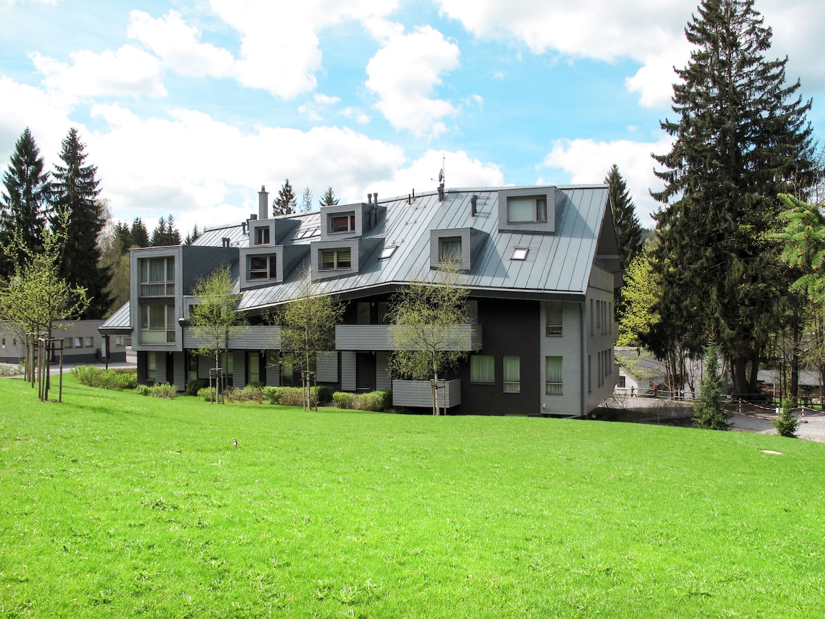 A modern residential building is set against a backdrop of greenery, featuring a combination of dark and light-colored facades. The surrounding grass is well-maintained, and several trees dot the landscape, adding to the natural ambiance of the area.