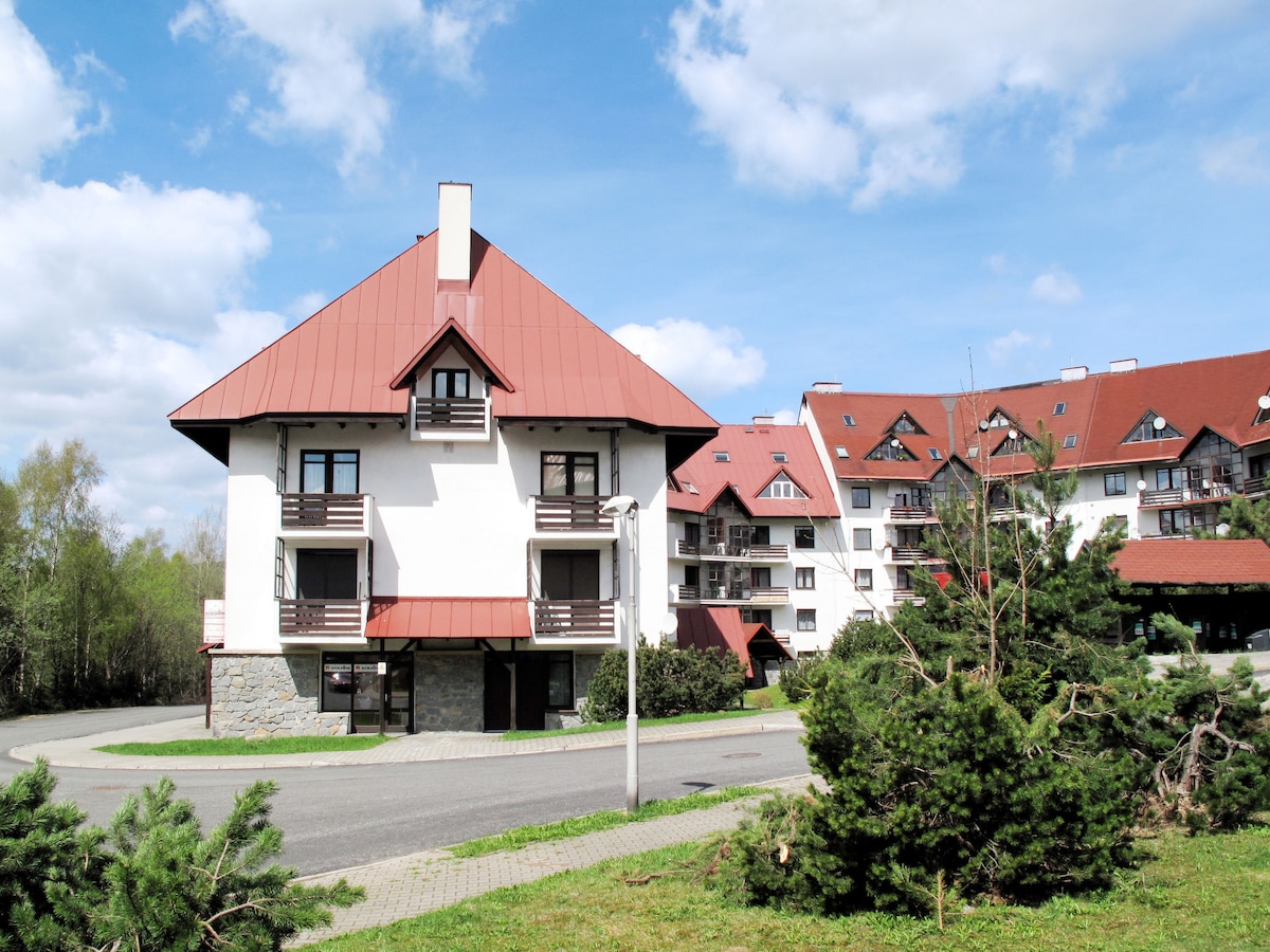A two-story building with a red sloped roof is situated in a green residential area. The exterior features multiple balconies and large windows. Surrounding trees and shrubs are visible, along with a well-maintained pathway leading to the entrance.
