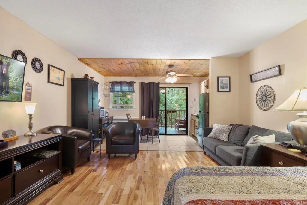 An open living area features polished wooden floors, a ceiling fan, and a sliding glass door leading to an outdoor deck. Comfortable seating options include two leather chairs and a spacious couch. A dining table for four is positioned next to a kitchenette with essential appliances.