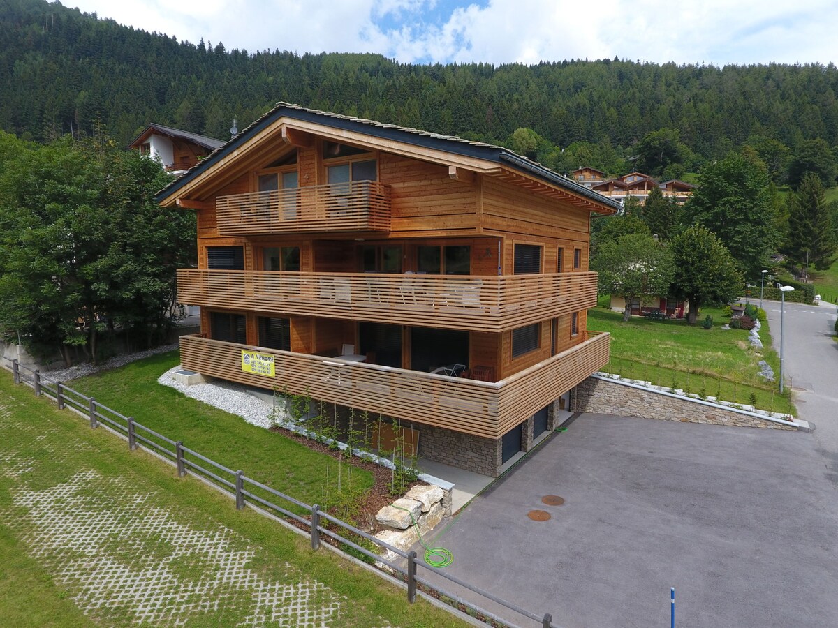 The exterior of a wooden chalet-style building is depicted, showcasing multiple balconies with natural wood detailing. A landscaped area with greenery surrounds the structure, and a paved parking area is visible in the foreground. The setting is framed by mountains and a clear sky.