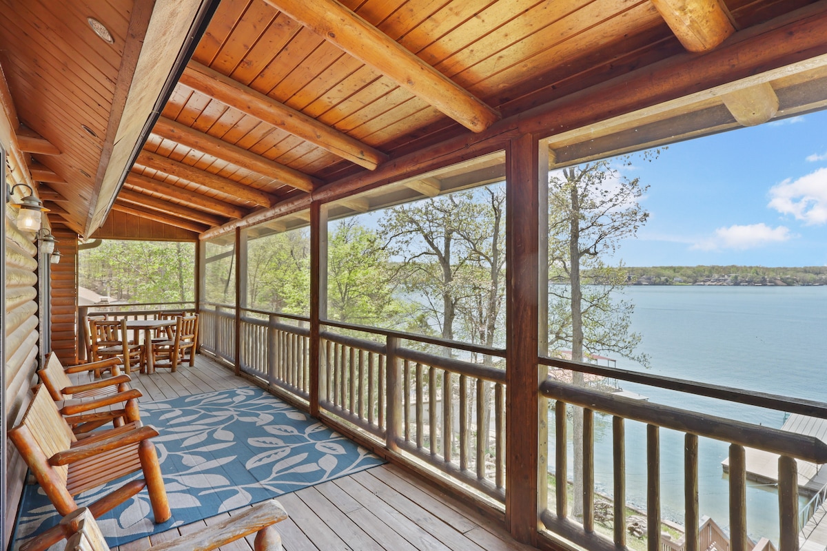 A spacious deck with wooden beams overlooks the serene lake. Comfortable seating options are available for relaxation, alongside a dining table for outdoor meals. The flooring is adorned with a patterned rug, and the natural surroundings can be enjoyed through the screened-in area.