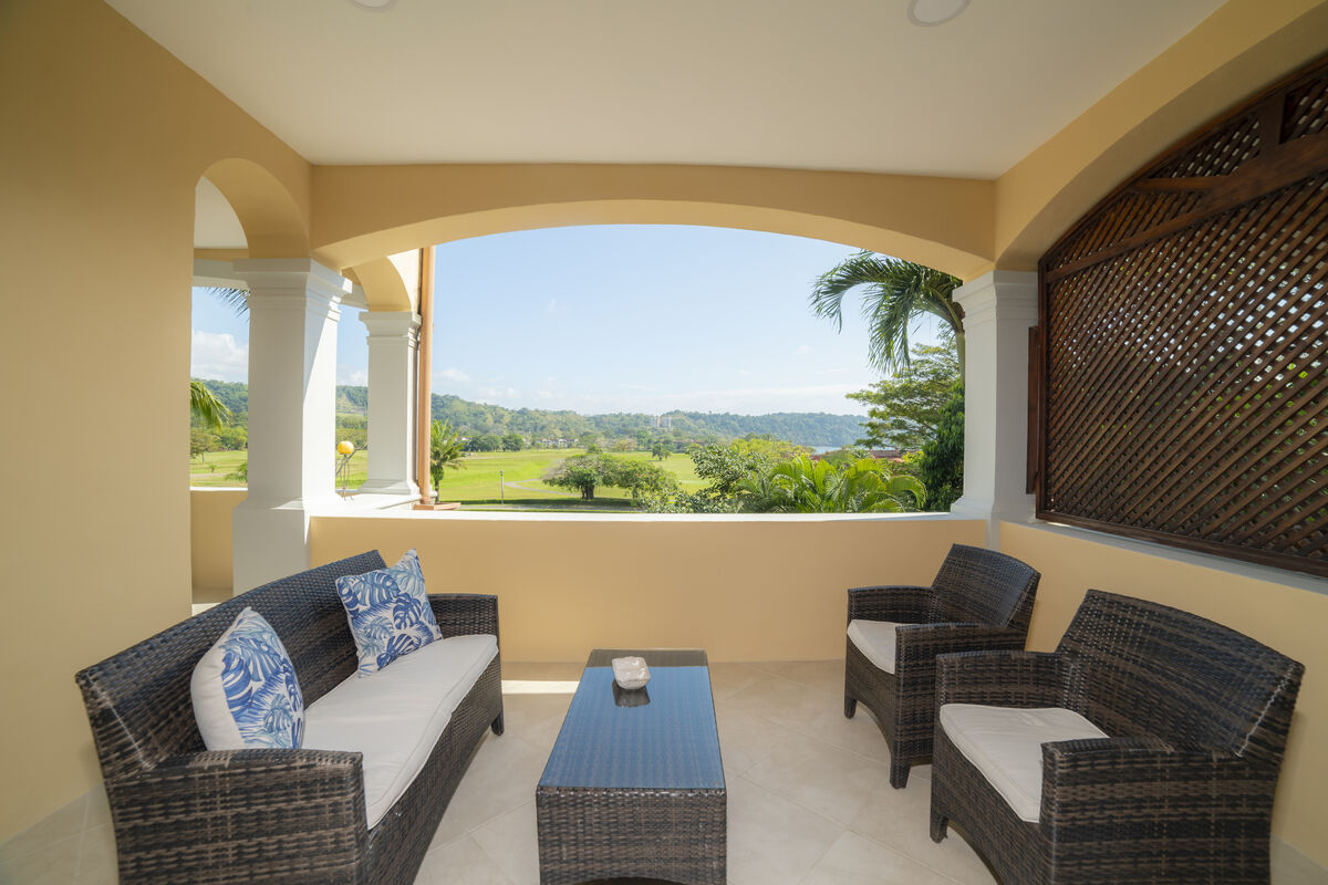 A covered terrace features a wicker seating arrangement comprising a sofa and two armchairs, complemented by decorative cushions. A rectangular table sits in the center. Expansive views of the lush landscape and golf course are visible through the arched opening, enhancing the serene setting.