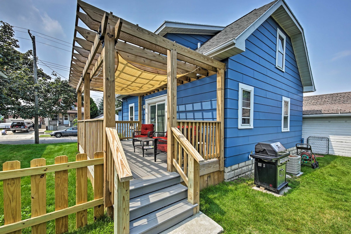 A welcoming outdoor deck is situated at the front of the cozy blue house, featuring shaded seating with red cushions. A picnic table is available for al fresco dining, and a gas grill is positioned nearby for seasonal barbecues. Lush green grass surrounds the area.