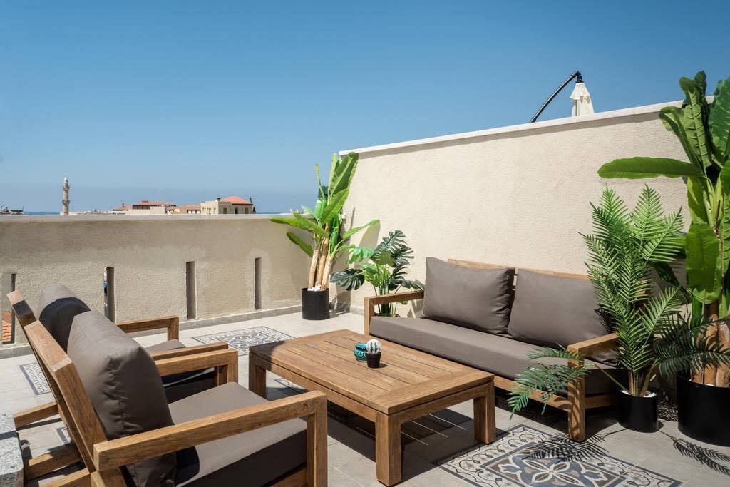 An outdoor terrace is furnished with wooden seating arrangements, featuring a coffee table and cushioned chairs. Lush greenery in planters adds a refreshing touch. The terrace showcases unobstructed views of the surrounding landscape under a clear blue sky.
