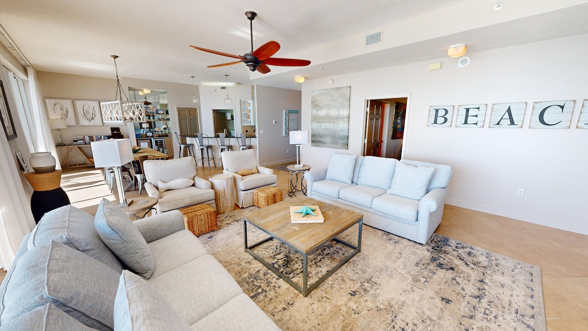 A spacious living area is furnished with light grey sofas and accent chairs, arranged around a central coffee table. A patterned area rug covers the floor, while large windows allow natural light to brighten the space. Decorative wall art reading 'BEACH' adds character to the room.