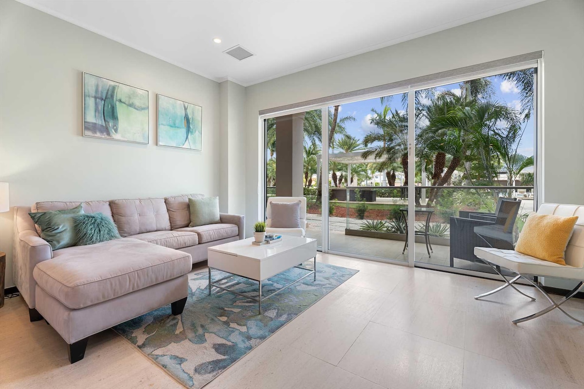 The living area is furnished with a light-colored sofa and an indoor coffee table, accentuated by a decorative rug. Large glass doors allow natural light to fill the space and offer views of lush tropical landscaping outside.