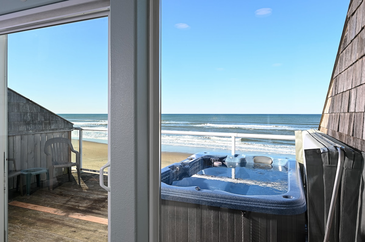 A private hot tub is positioned on a spacious oceanfront deck, offering stunning views of the beach and waves. Light blue water contrasts with the sandy shore, while two white outdoor chairs invite relaxation under clear blue skies.