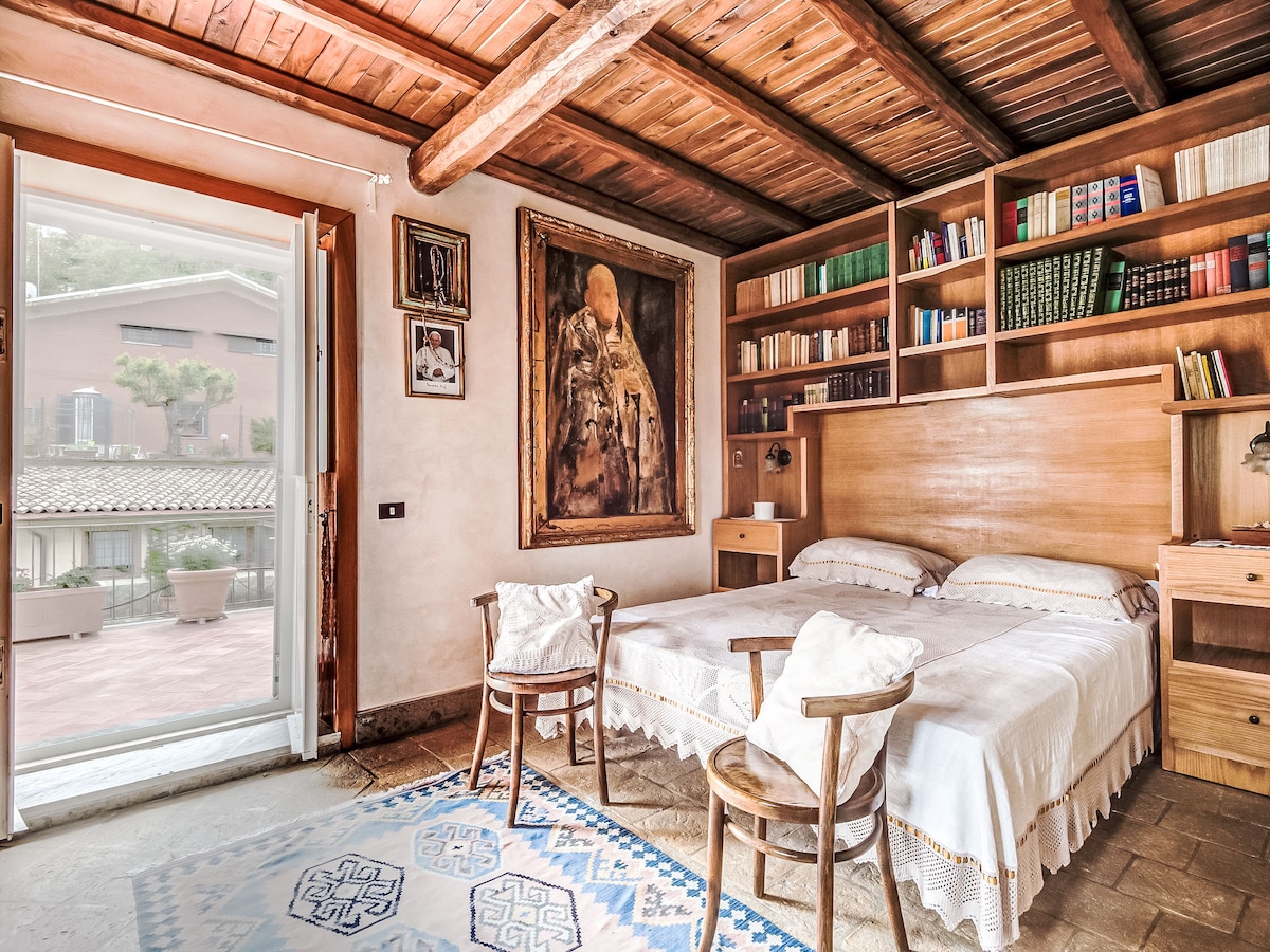 A warm bedroom is presented with wooden beams and a rustic ceiling. The space includes a spacious bed with a light coverlet, flanked by wooden bookshelves filled with books. A large artwork adorns one wall, and a door opens to a balcony with garden views.