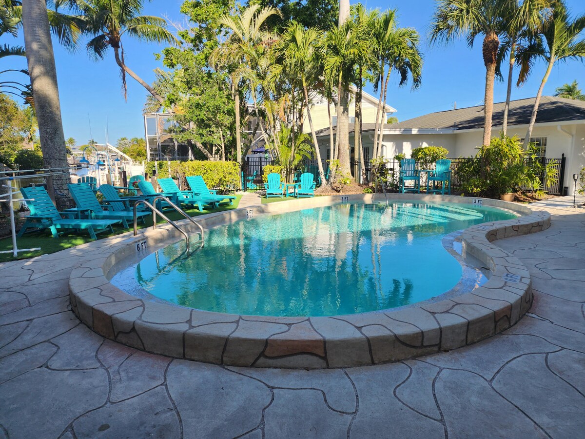 A communal pool is framed by palm trees, with lounge chairs positioned around its edges. The water reflects the sky, creating a serene setting. The stone pool deck is surrounded by vibrant greenery, enhancing the natural ambiance of the area.
