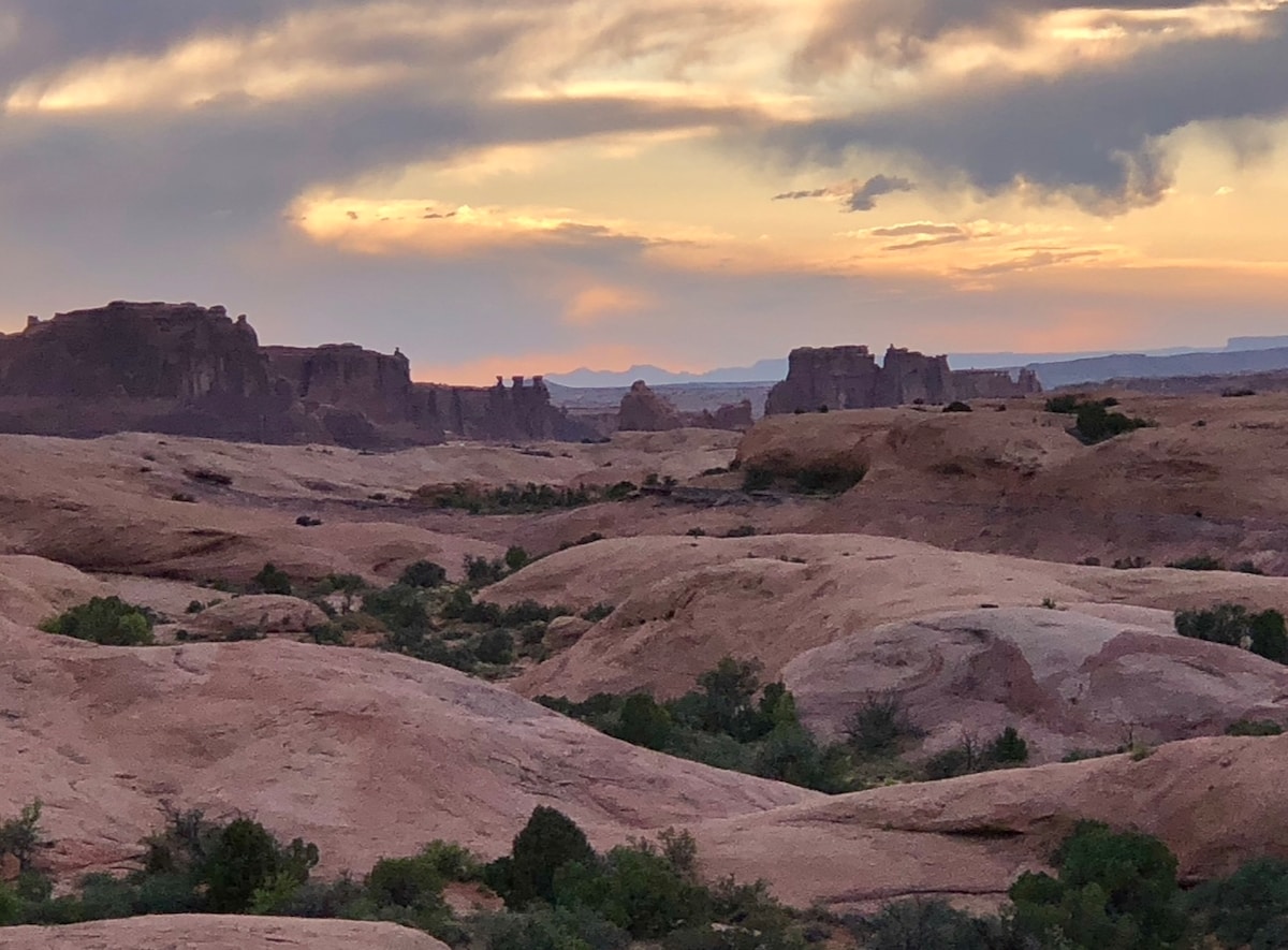 A vast desert landscape is showcased, featuring rolling hills of reddish-brown earth dotted with patches of green vegetation. The sky is filled with soft, muted hues of purple and orange as the sun sets, illuminating the distant rock formations that rise dramatically against the horizon.