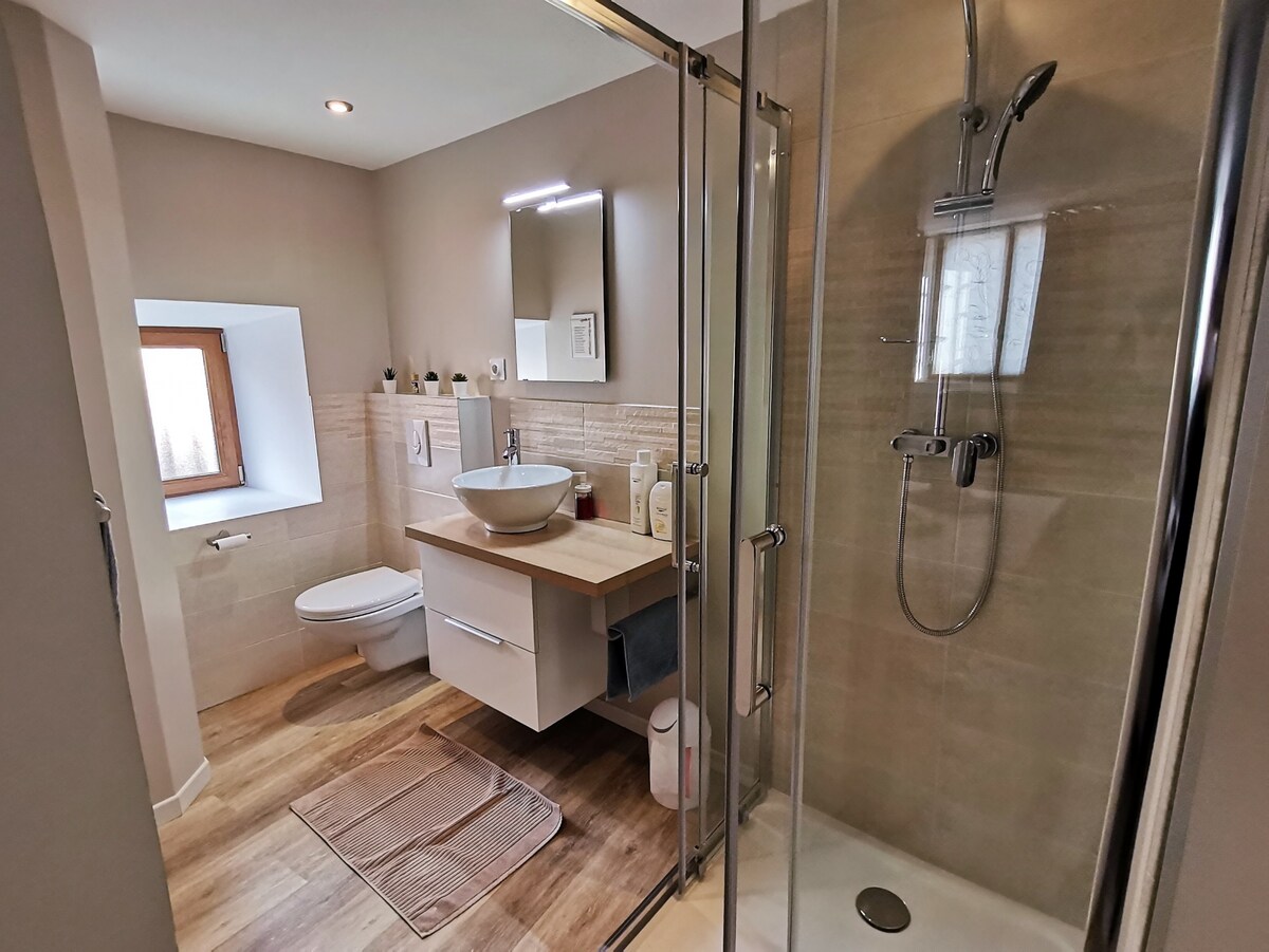 The modern bathroom features a walk-in shower with a glass enclosure and a sleek rainfall showerhead. A floating vanity with a round basin is complemented by essential toiletries. Natural light filters through a small window, illuminating the neutral-toned tiles and warm wooden floor.