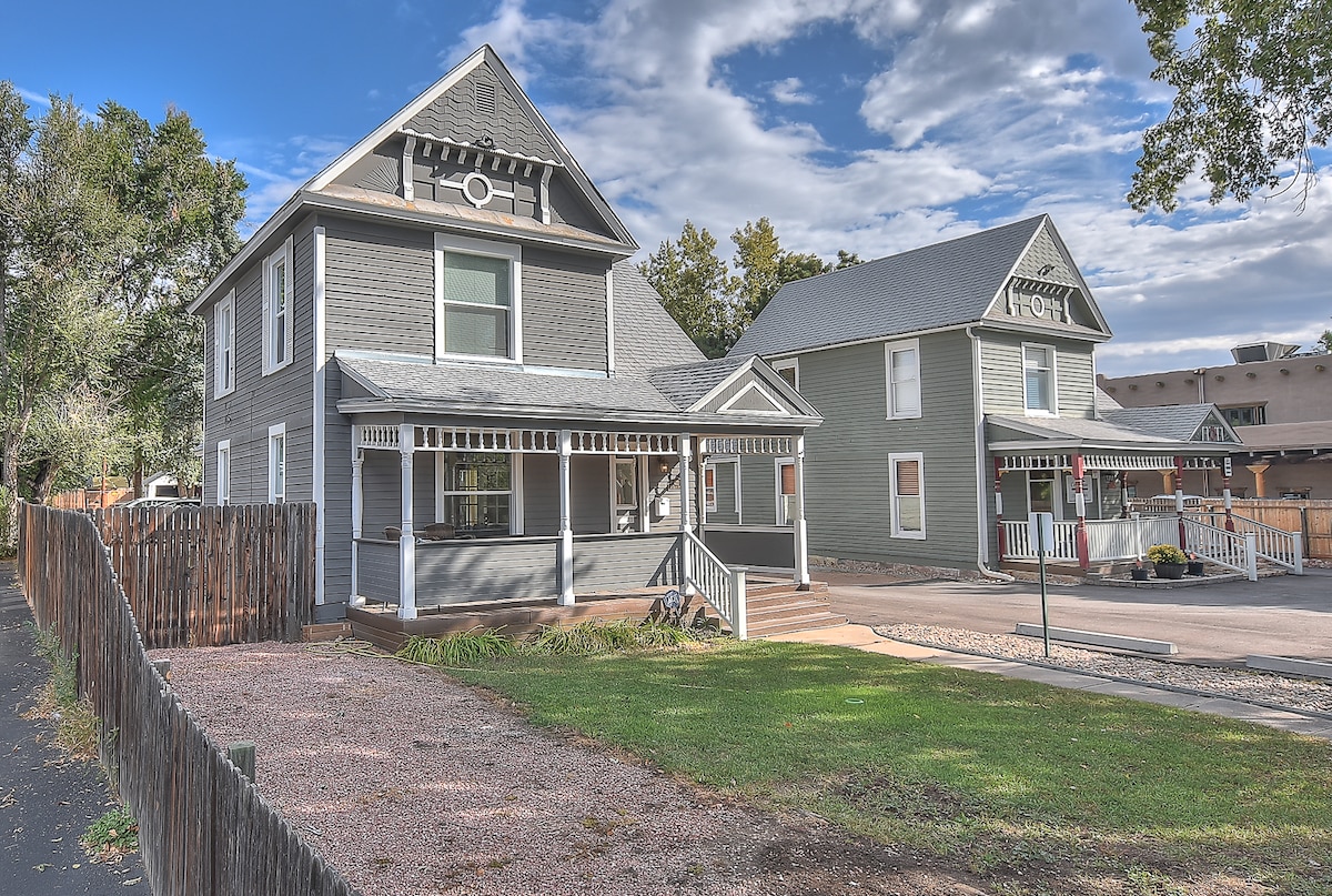 A Victorian-style house is depicted, featuring a covered front porch with white railing and steps leading to a welcoming entrance. The exterior showcases gray siding and is surrounded by a well-maintained lawn. Nearby, another house is visible, contributing to the neighborhood's character.