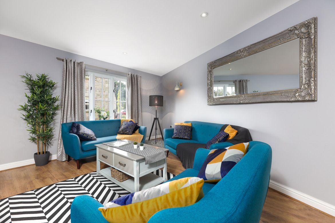 A welcoming living area is showcased, featuring a mix of blue and black sofas complemented by patterned cushions. A glass-topped coffee table sits on a striped rug, and natural light filters through the large windows, with curtains framing the view. A decorative mirror adds depth to the space.