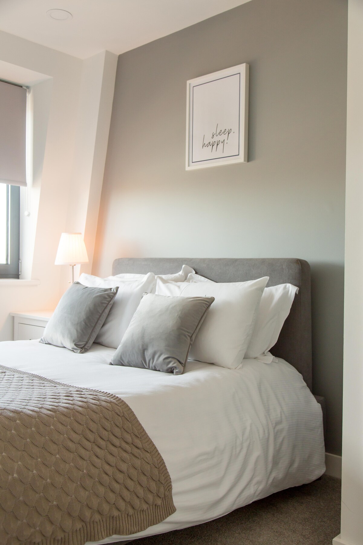 A softly lit bedroom features a spacious bed adorned with plush grey and white pillows. A textured blanket drapes elegantly over the bed, while a framed print on the wall adds a touch of personalization. Natural light enters through a window fitted with a roller shade.