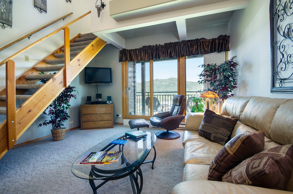 The living area features a comfortable leather sofa arranged around a glass coffee table, accompanied by a cozy armchair. A mounted television is positioned above a wooden cabinet. Large windows frame scenic views of the outdoor landscape and provide access to the private balcony.