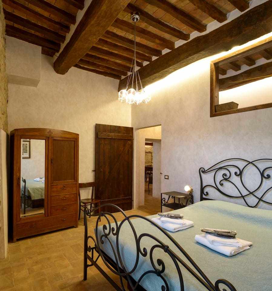 A spacious bedroom features a wrought-iron bed dressed in soft linens. Natural wooden beams accentuate the ceiling. A wooden dresser and a mirror provide practical elements. Soft lighting illuminates the room, creating a calm atmosphere. An arched wooden door leads to the adjoining living area.