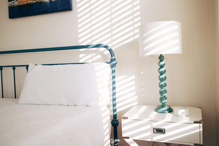 Un armazón de cama de metal azul, complementado con sábanas blancas, y una mesita de noche con una lámpara decorativa