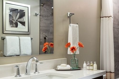 A well-appointed bathroom features a modern sink with sleek faucets and a large mirror reflecting the space. Two neatly arranged towels hang on the rack. A vase of orange flowers provides a pop of color on the countertop, alongside toiletry bottles and a folded washcloth.