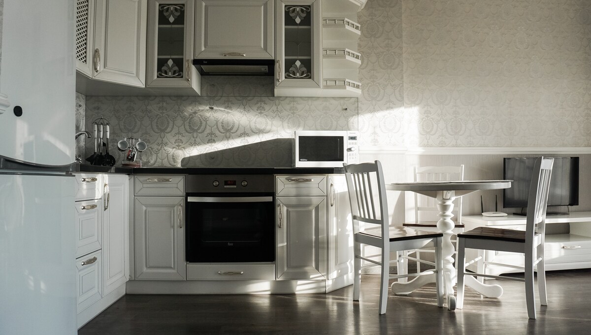 The kitchen area features white cabinetry and a modern design. An induction cooktop and oven are installed, along with a microwave and refrigerator. A small dining table with chairs is positioned in the center, illuminated by natural light streaming through the window.