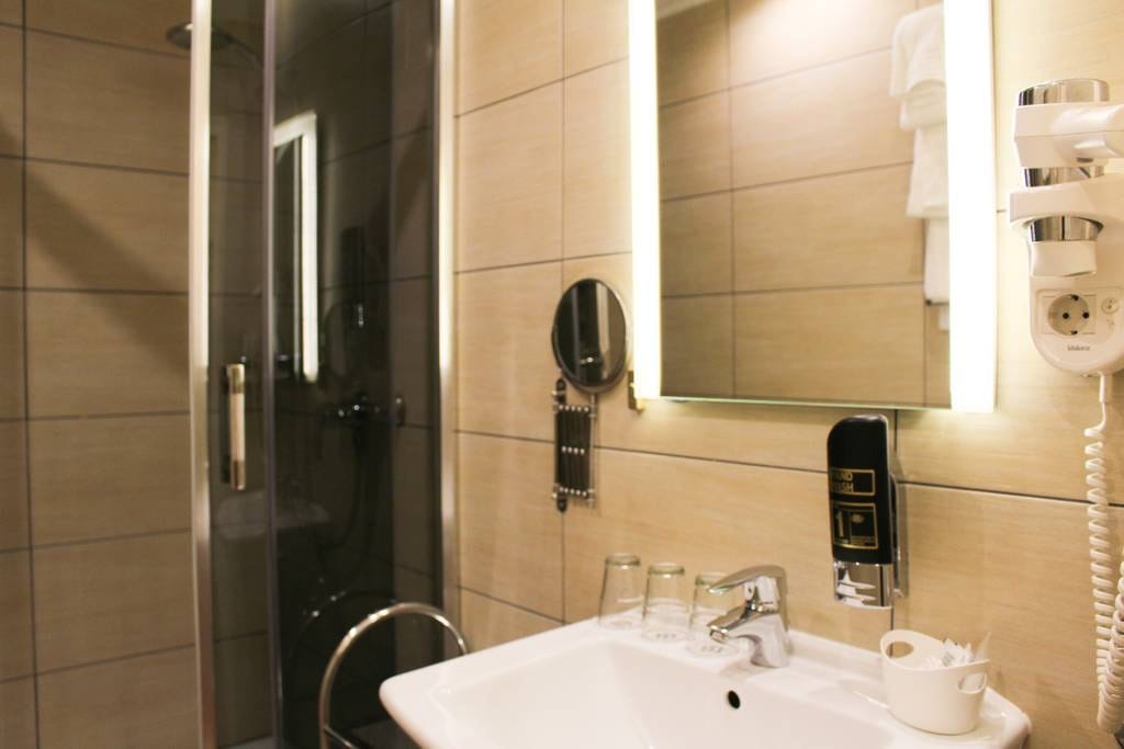 A modern bathroom is equipped with a walk-in shower, illuminated by a sleek mirror above a compact sink. Glasses and toiletries are neatly arranged, while a hairdryer and a round mirror are mounted on the wall, enhancing functionality.