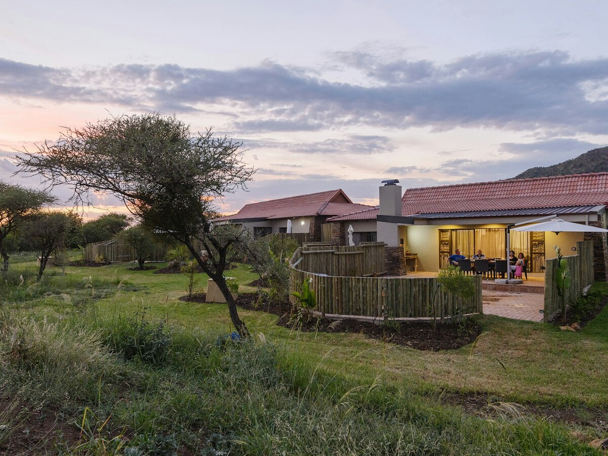 The exterior of the villa is showcased during twilight, featuring a patio area with seating and a dining table under a shaded structure. Surrounding greenery includes scattered trees and lush grass, enhancing the natural setting against a backdrop of soft, colorful clouds.