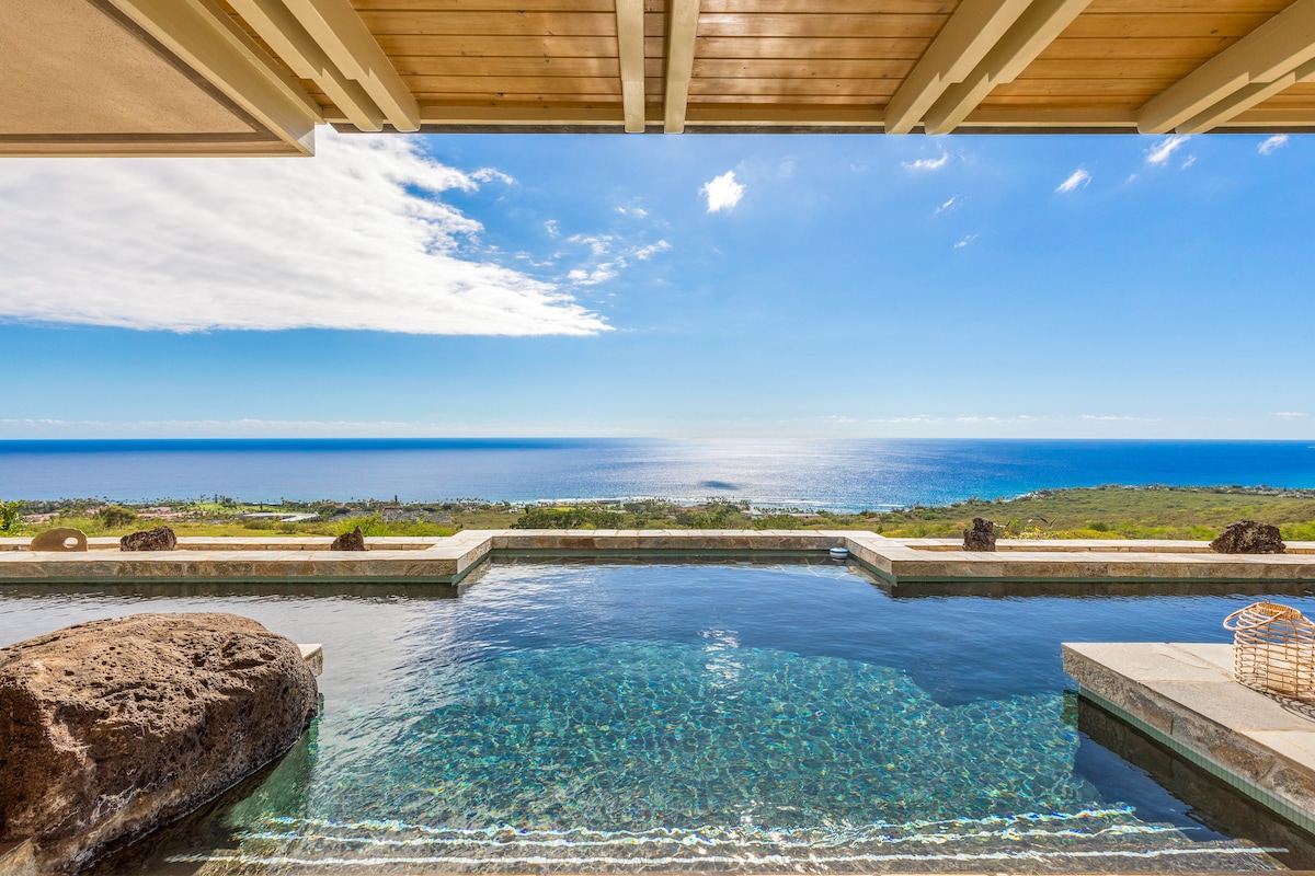 An infinity pool stretches toward the horizon, showcasing expansive ocean views under a clear blue sky. The tranquil water reflects the surrounding landscape, with natural stone elements integrated into the pool design, creating a harmonious connection between the home and nature.