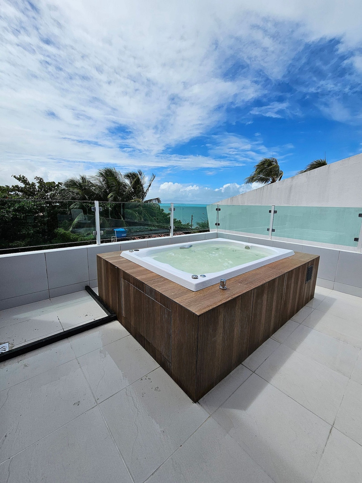A hot tub is situated on a spacious terrace, surrounded by glass railings that provide unobstructed views of the landscape. The surface is tiled and reflects natural light, while the sky above offers a mix of blue and white clouds.