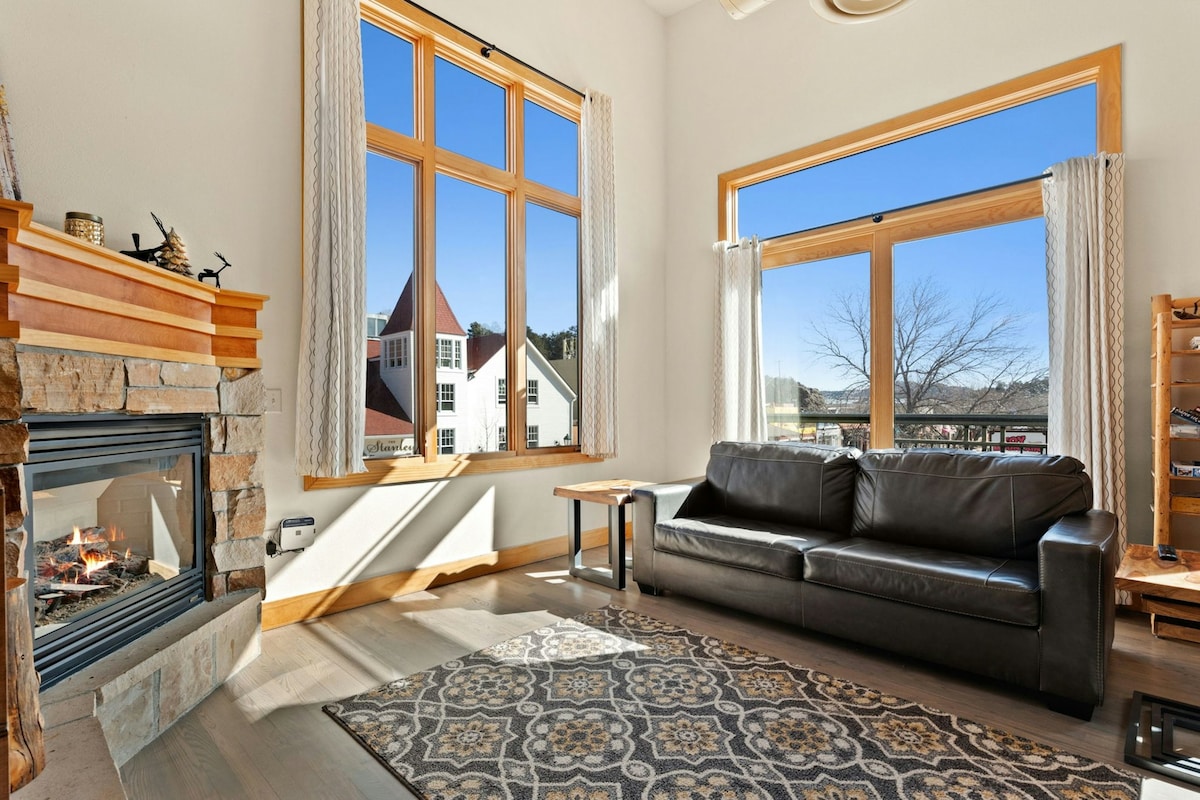 A bright living room is highlighted by large windows that overlook the scenic surroundings. A sleek black sofa is positioned next to a stone fireplace, providing warmth and comfort. A decorative area rug adds texture to the hardwood floor, enhancing the cozy ambiance.