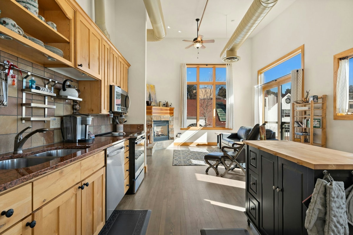 The kitchen area features wooden cabinetry and stainless steel appliances, complemented by a large island with seating. Natural light fills the open space, highlighting a comfortable living area with a fireplace and large windows, revealing views of the outdoors.
