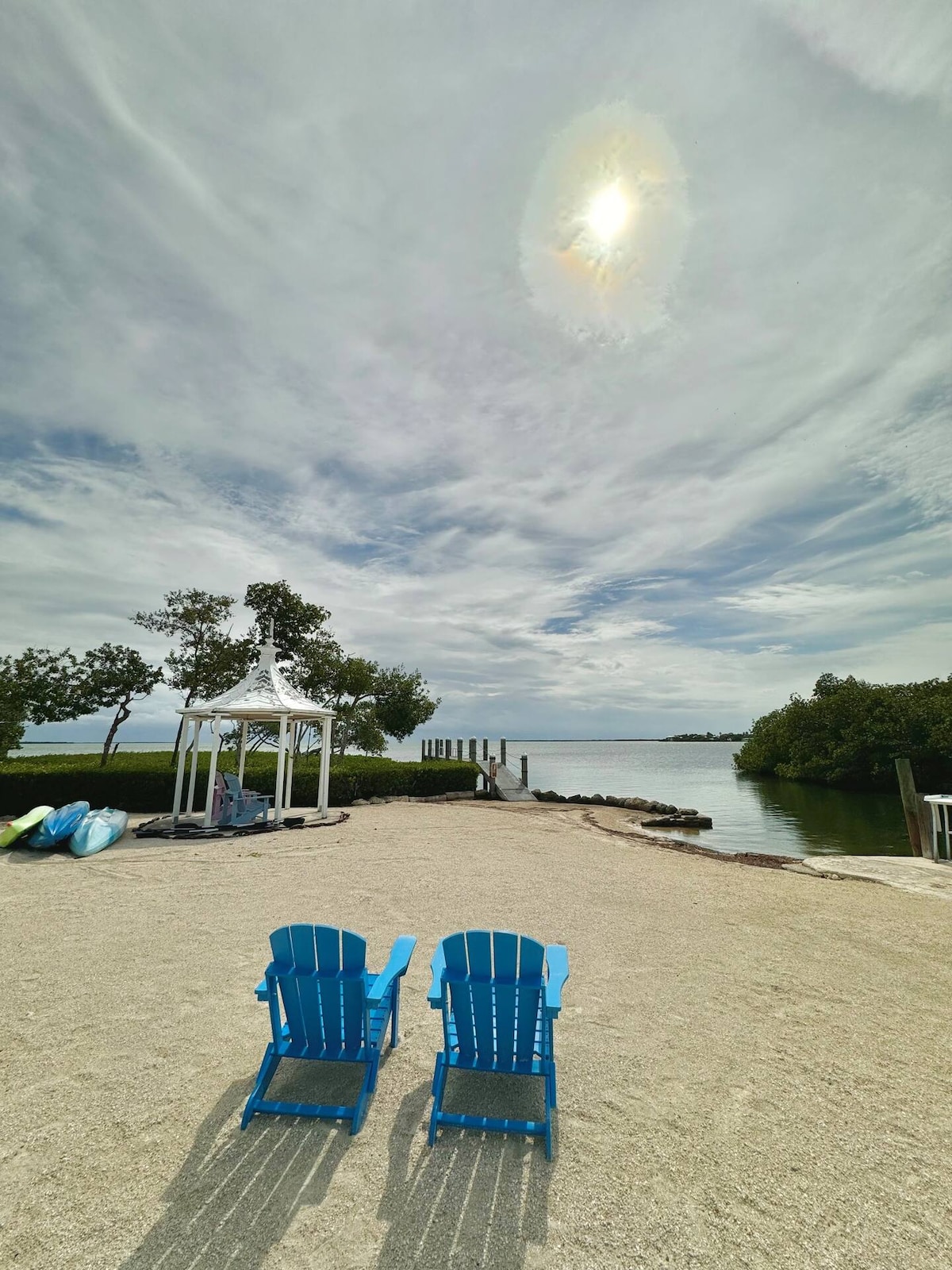 Two bright blue Adirondack chairs are positioned on sandy ground, facing a serene waterway. A gazebo is visible in the background, surrounded by trees. Kayaks are aligned to the left, while a wooden pier extends into the tranquil water, reflecting the cloudy sky above.