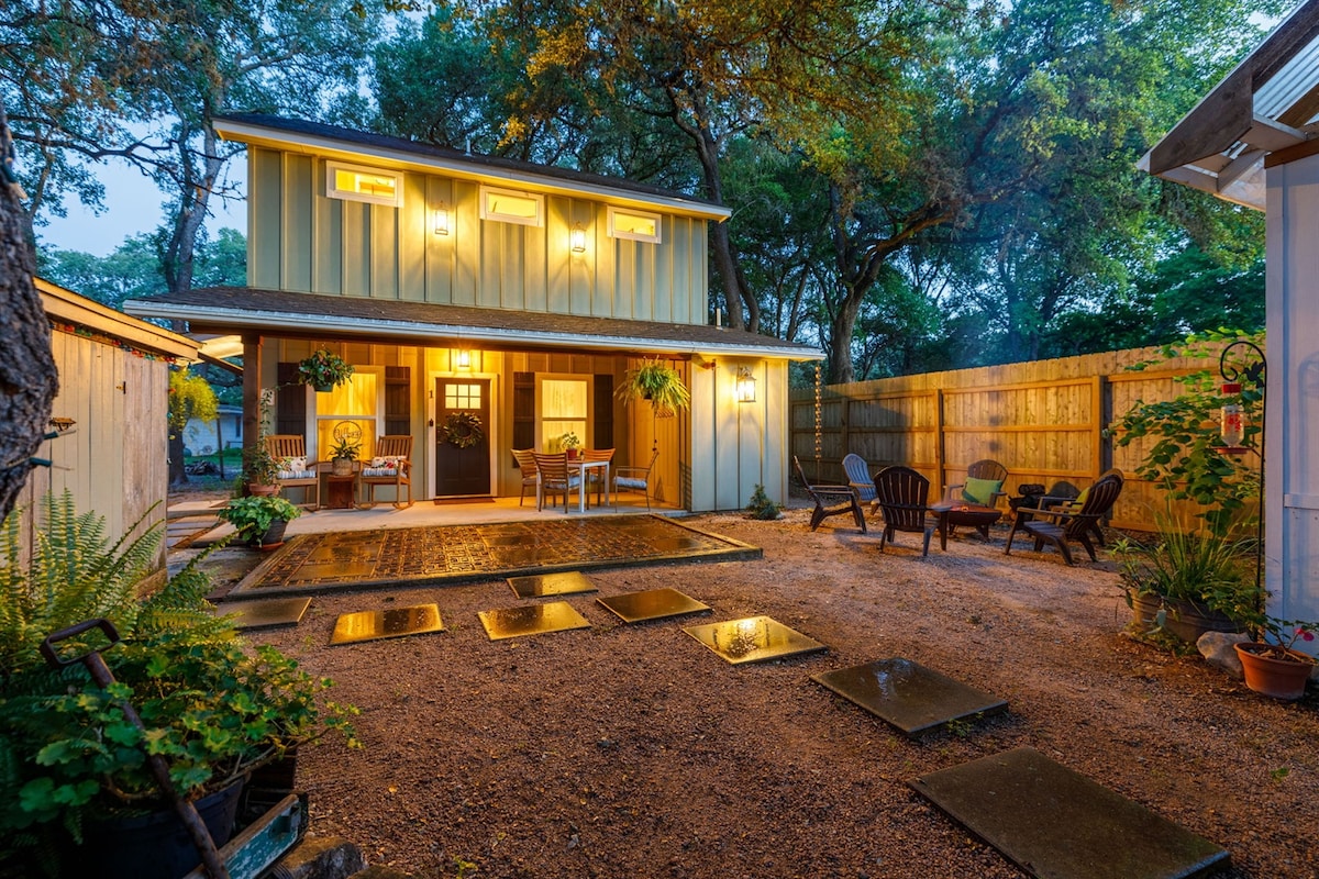 The outdoor area showcases a gravel path leading to the guest house entrance, flanked by potted plants. Seating arrangements include two chairs on the porch and several outdoor chairs arranged around a fire pit, creating a welcoming space for relaxation.