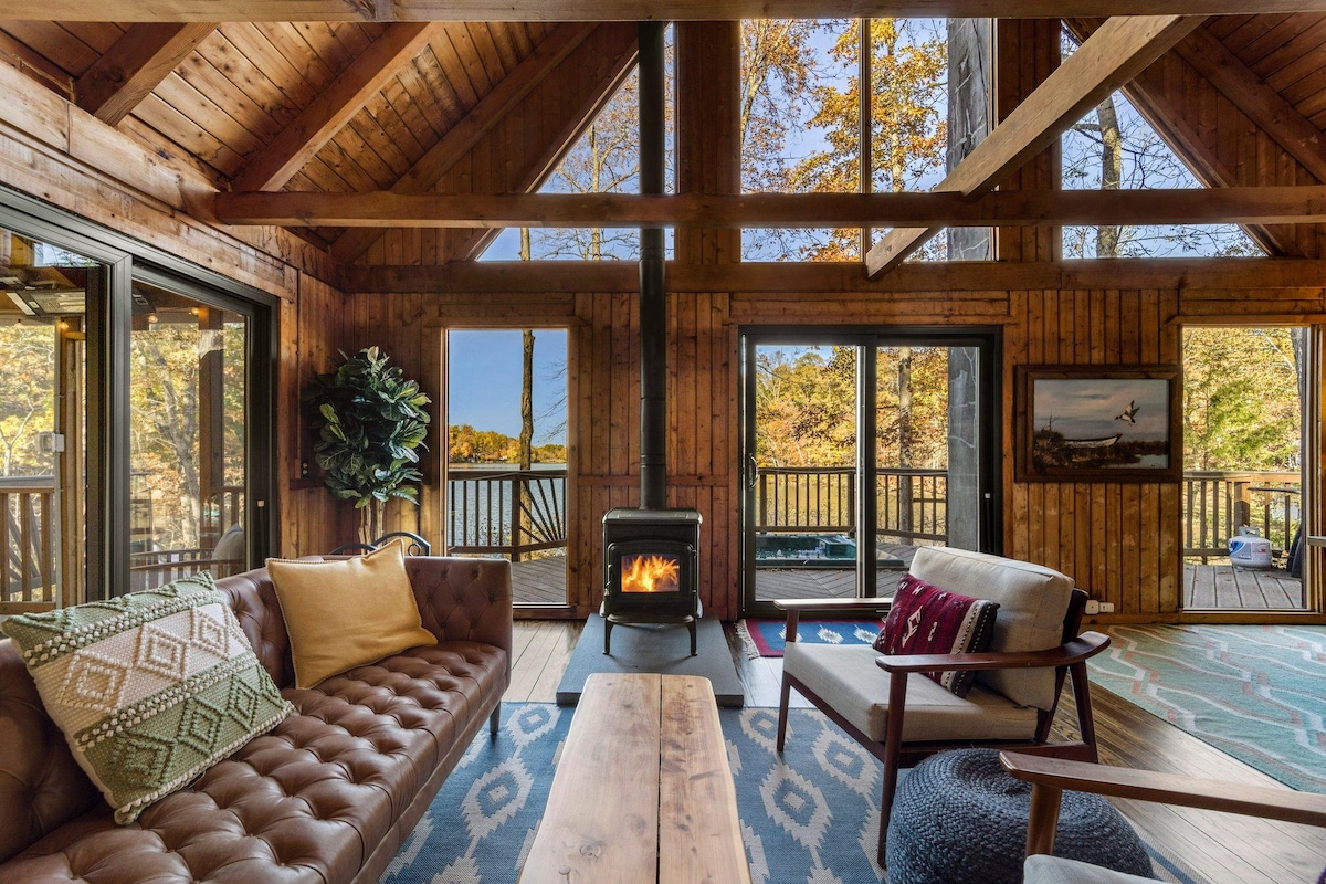 The living area is designed with high, exposed wooden beams and large windows framing lake views. A cozy wood stove is positioned near a leather sofa, set against warm wooden walls. A natural wood coffee table sits in the center, complementing the surrounding seating.