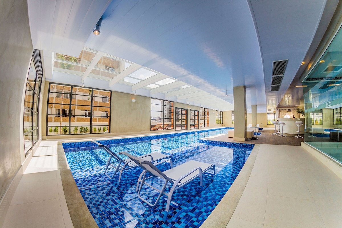 An indoor pool area is highlighted by a bright, open design. Sunlight filters through large glass panels above, illuminating the blue-tiled pool. Several lounge chairs are situated on the pool deck, with a small seating area visible in the background.