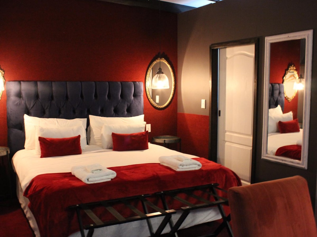 A cozy sleeping area features a king-size bed with a deep blue tufted headboard, complemented by decorative pillows in rich red. A mirror and two lamps add elegance, while modern furnishings are arranged to enhance comfort. Natural light filters through an adjacent door.