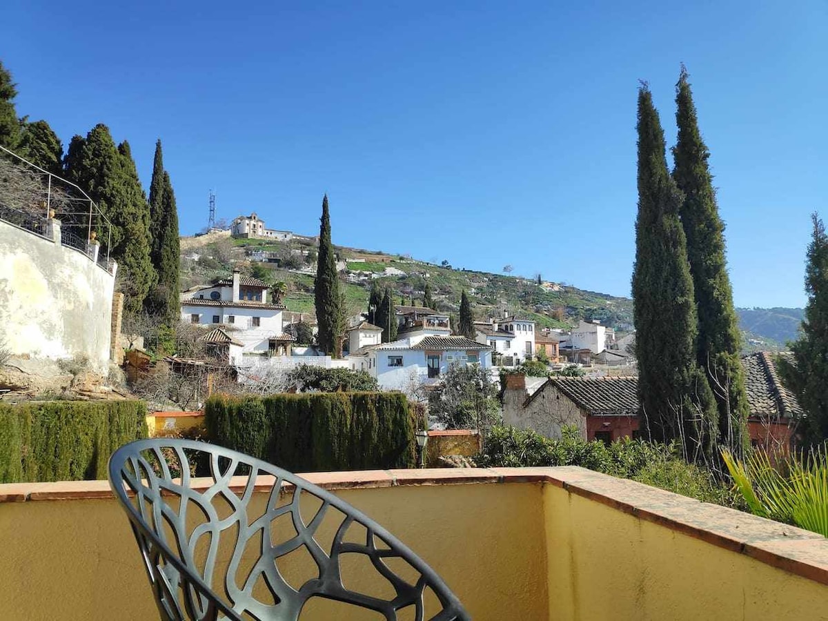 A serene outdoor view showcases lush greenery and distant hills, framed by tall cypress trees. The scene includes traditional whitewashed houses nestled in the landscape, under a clear blue sky.