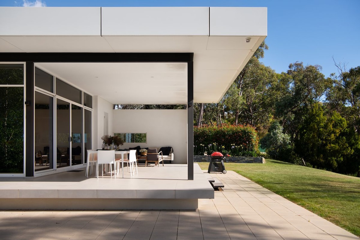 An outdoor view of a contemporary home’s patio area is presented, featuring a spacious concrete platform with a dining set for six. Large glass doors open to the interior, allowing natural light to flood the space, while a barbecue grill is positioned on the lawn.