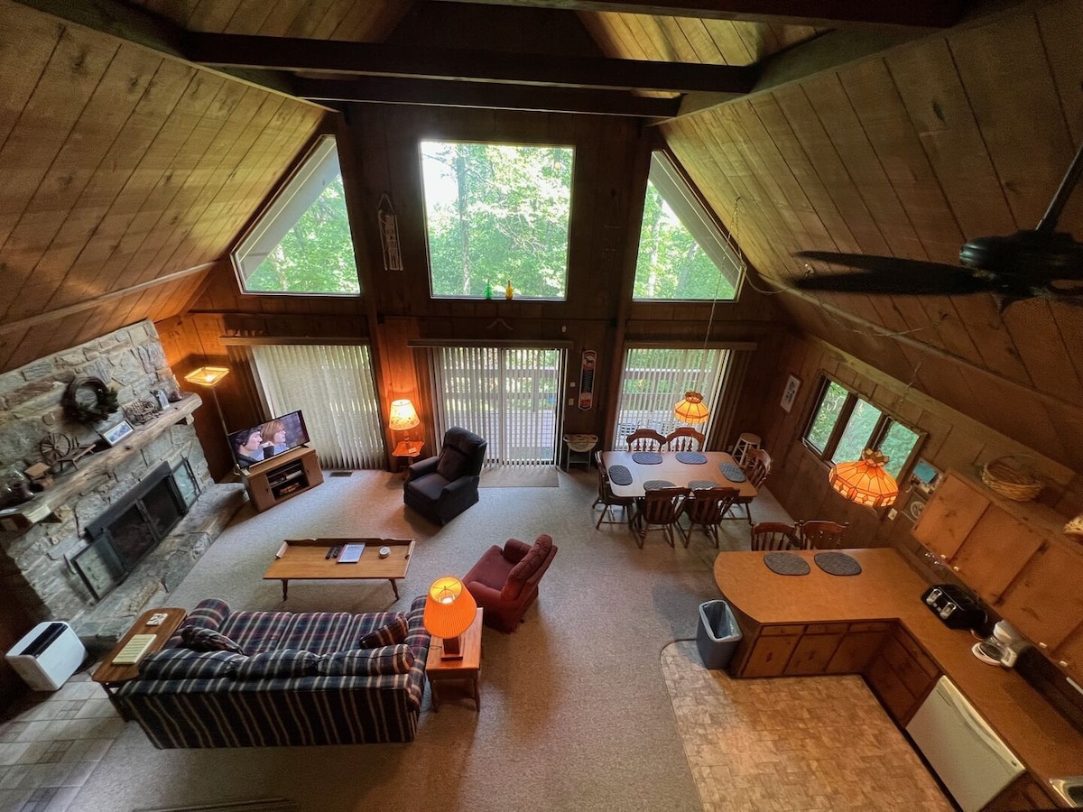 The great room features a stone fireplace and ample seating, including a plaid couch and two armchairs. Large windows allow natural light to fill the space. A dining area with a wooden table and six chairs is visible, along with a well-equipped kitchen area.