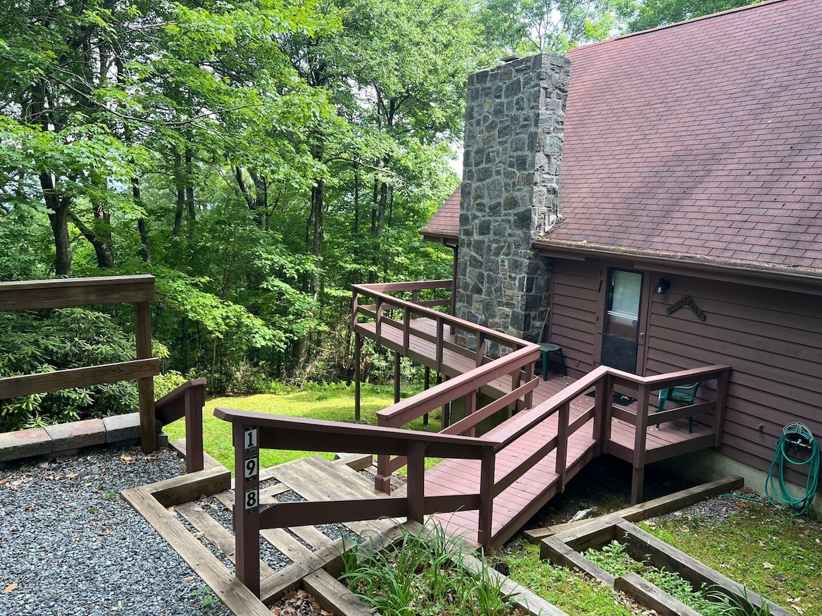 A stone fireplace is featured on the side of the chalet, with a gradual ramp leading to the entrance. The surrounding trees create a natural backdrop, while gravel pathways and wooden decking provide access. The exterior showcases a rustic design with a pitched roof.