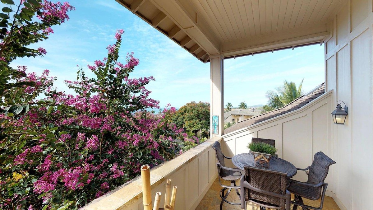 A private lanai offers a serene space adorned with a round table surrounded by dark wicker chairs. Lush pink flowering plants frame the view, creating a vibrant connection to the outdoors. The warm sunlight enhances the tranquil atmosphere of this outdoor retreat.