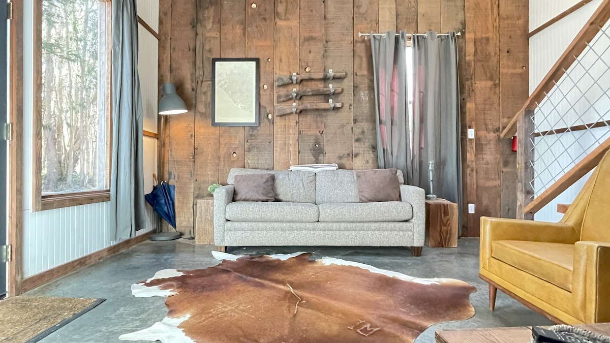 A warm living area features a textured wood accent wall and a cozy grey sofa, complemented by a stylish yellow armchair. Natural light filters through large windows, offering views of the surrounding trees. A decorative rug enhances the rustic decor.